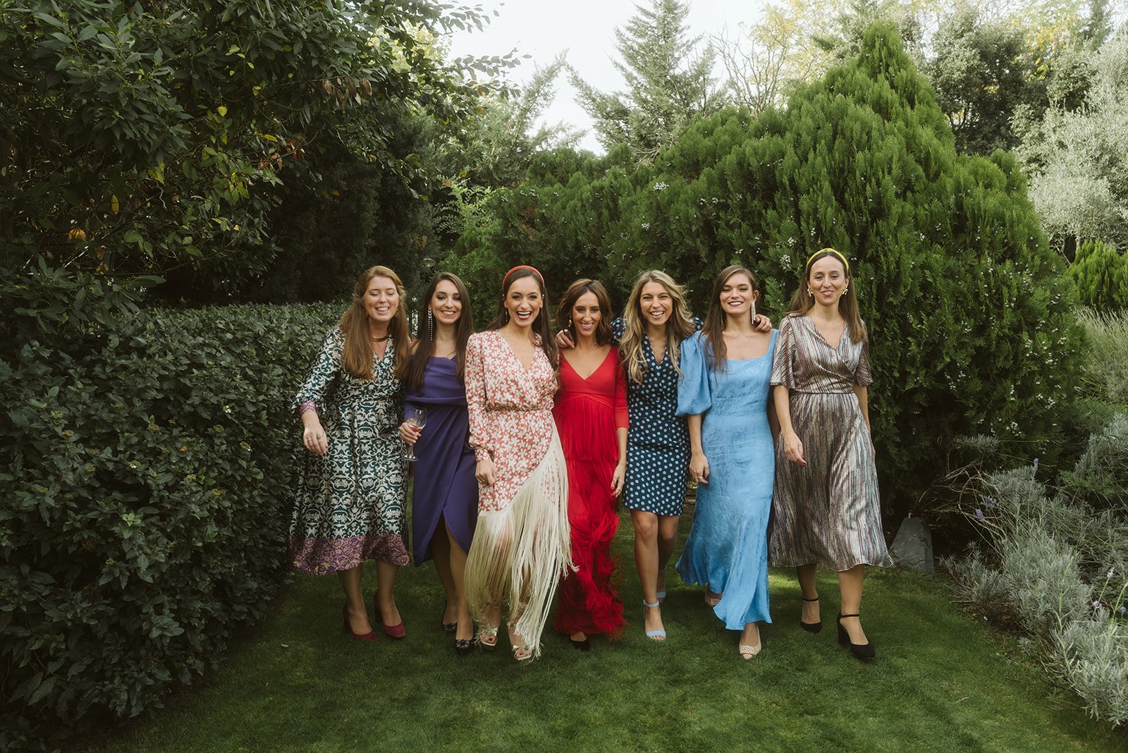 Siete mujeres posando sonrientes en un jardín, vistiendo vestidos coloridos y elegantes. Están rodeadas de vegetación exuberante.