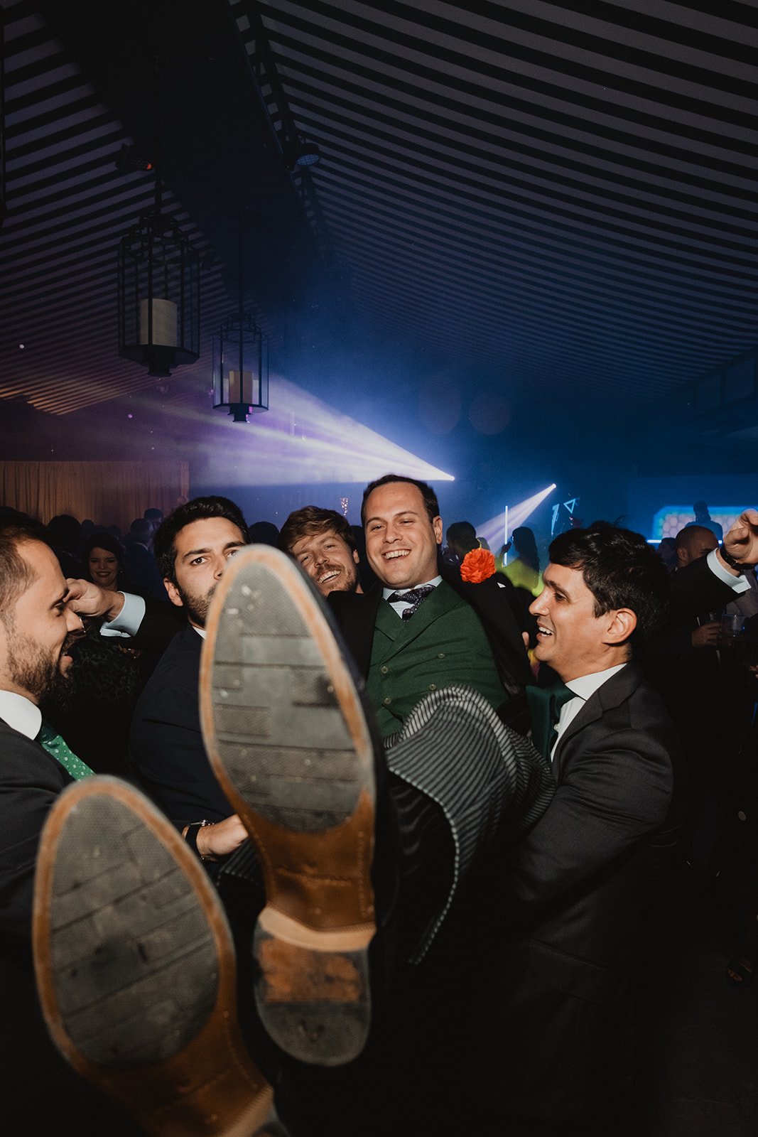 Grupo de hombres en traje levantando a otro en una celebración, bajo luces de fiesta.