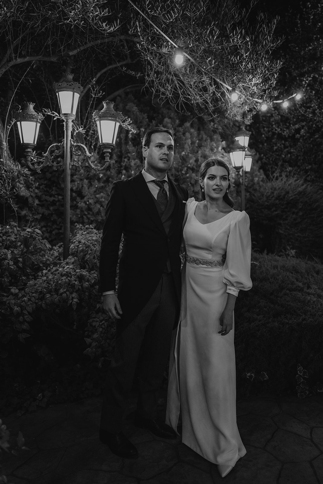 Pareja vestida formalmente en una escena nocturna con luces y vegetación de fondo.