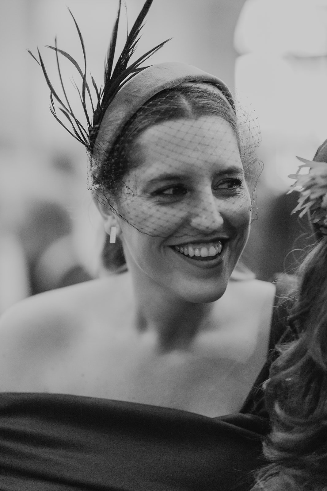 Una mujer sonriente con un fascinator con plumas y una malla en el cabello en una imagen en blanco y negro.
