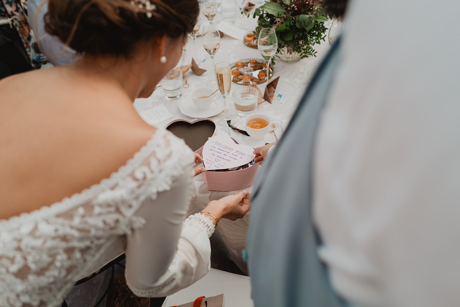 Mujer con vestido blanco abre una caja en forma de corazón durante una celebración, posiblemente una boda, en una mesa con copas y flores.