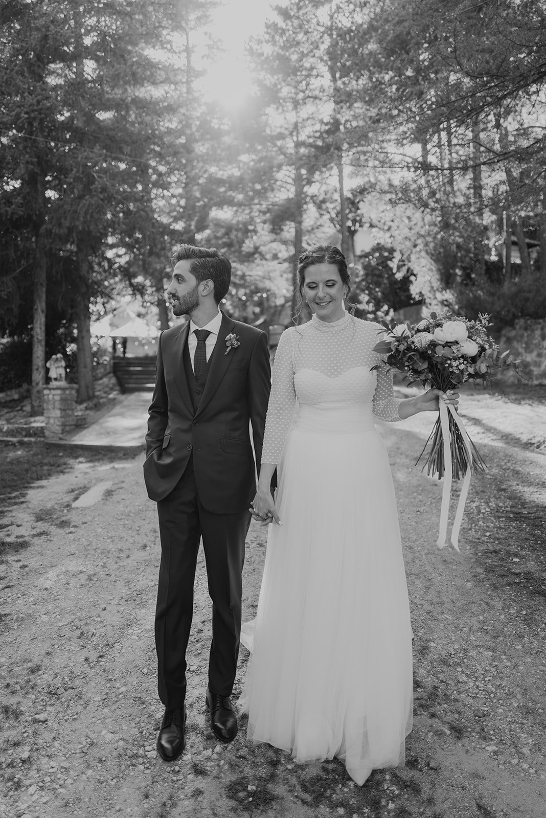 Pareja de novios caminando por un camino arbolado, ella sostiene un ramo de flores.