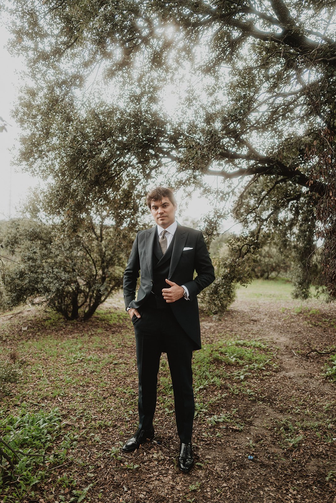 Hombre de pie con traje elegante en un entorno natural, rodeado de árboles y vegetación.