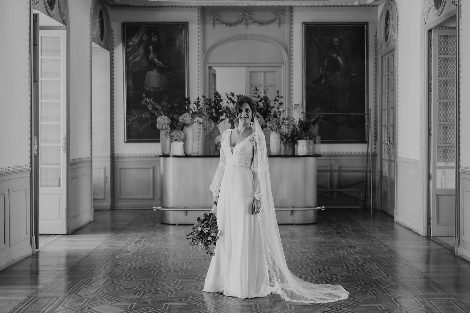 Novia en vestido de boda blanco, con ramo de flores, posando en un salón elegante con cuadros y decoración clásica.