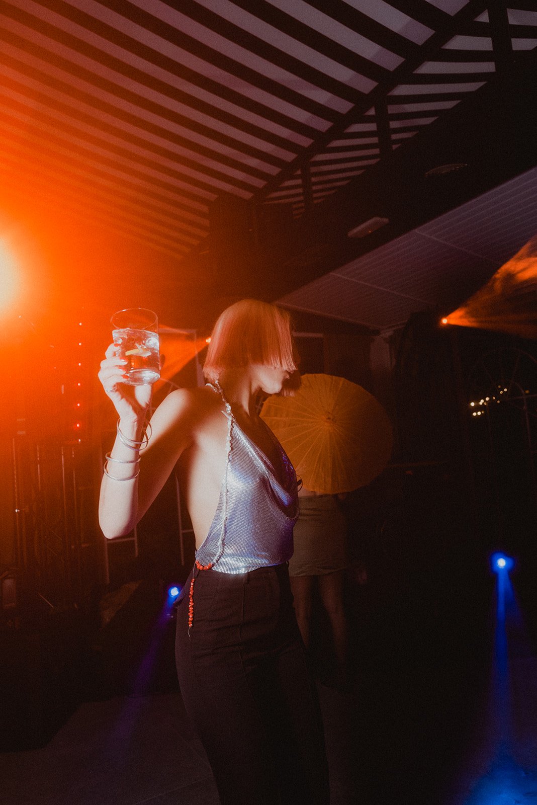 Persona con cabello corto rubio bailando en una fiesta, con luz roja intensa, vistiendo un top plateado y sosteniendo un vaso.