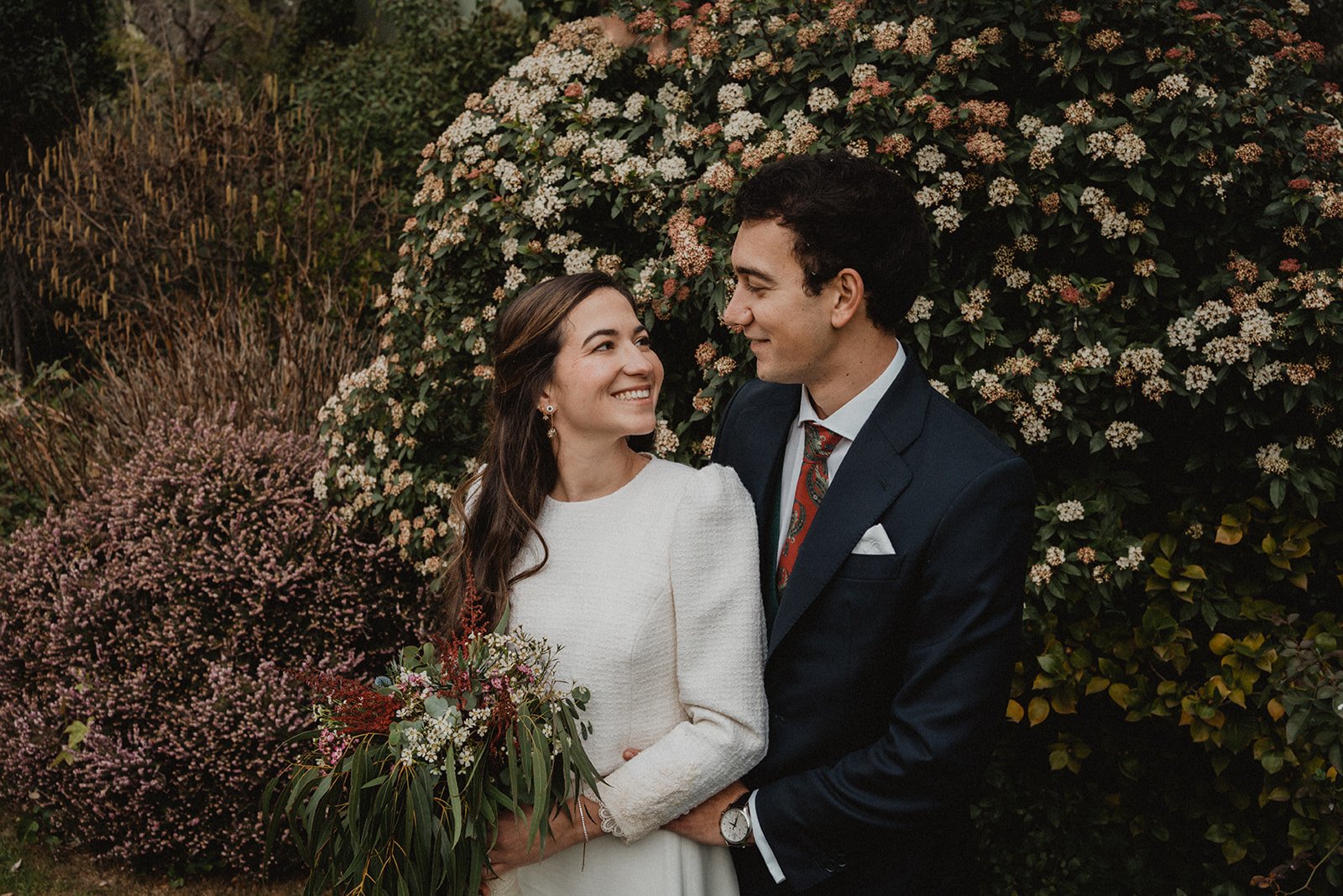 Pareja sonriente vestida elegantemente, ella con vestido blanco y ramo de flores, él con traje oscuro, rodeados de vegetación y flores.