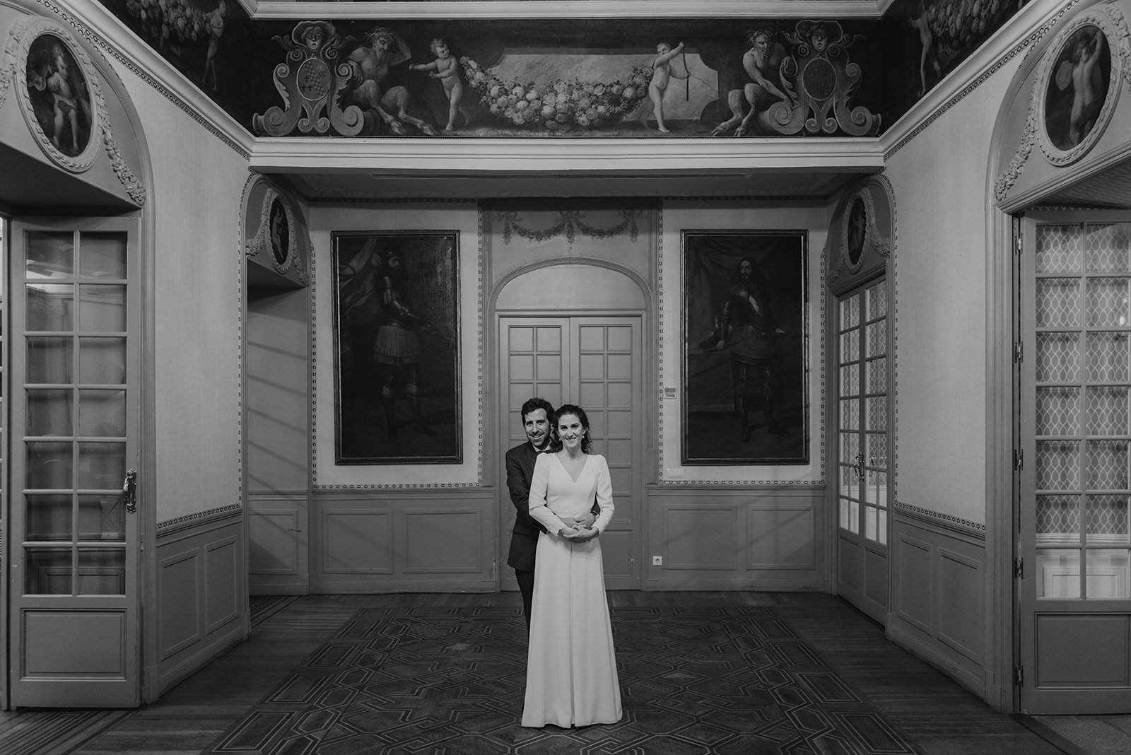 Pareja posando en una habitación elegante con pinturas en las paredes, puertas decorativas y detalles arquitectónicos clásicos en blanco y negro.