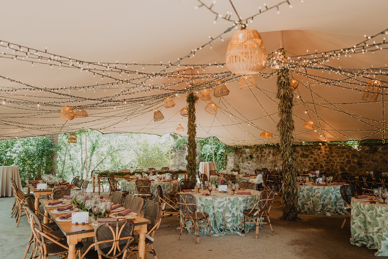 Interior de una carpa decorada para un evento, con mesas cubiertas con manteles de hojas verdes, sillas de mimbre, luces colgantes y lámparas de mimbre. Ambiente rústico y festivo, con paredes de piedra y vegetación alrededor.