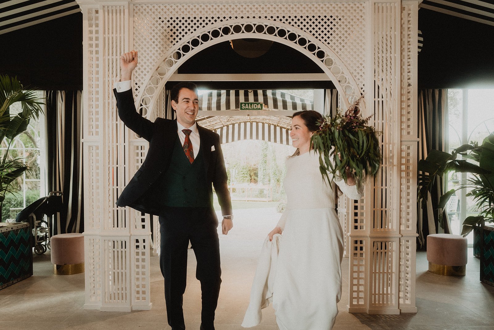 Una pareja vestida de forma elegante celebrando en un lugar decorado con plantas y estructuras blancas. La mujer lleva un ramo de flores.