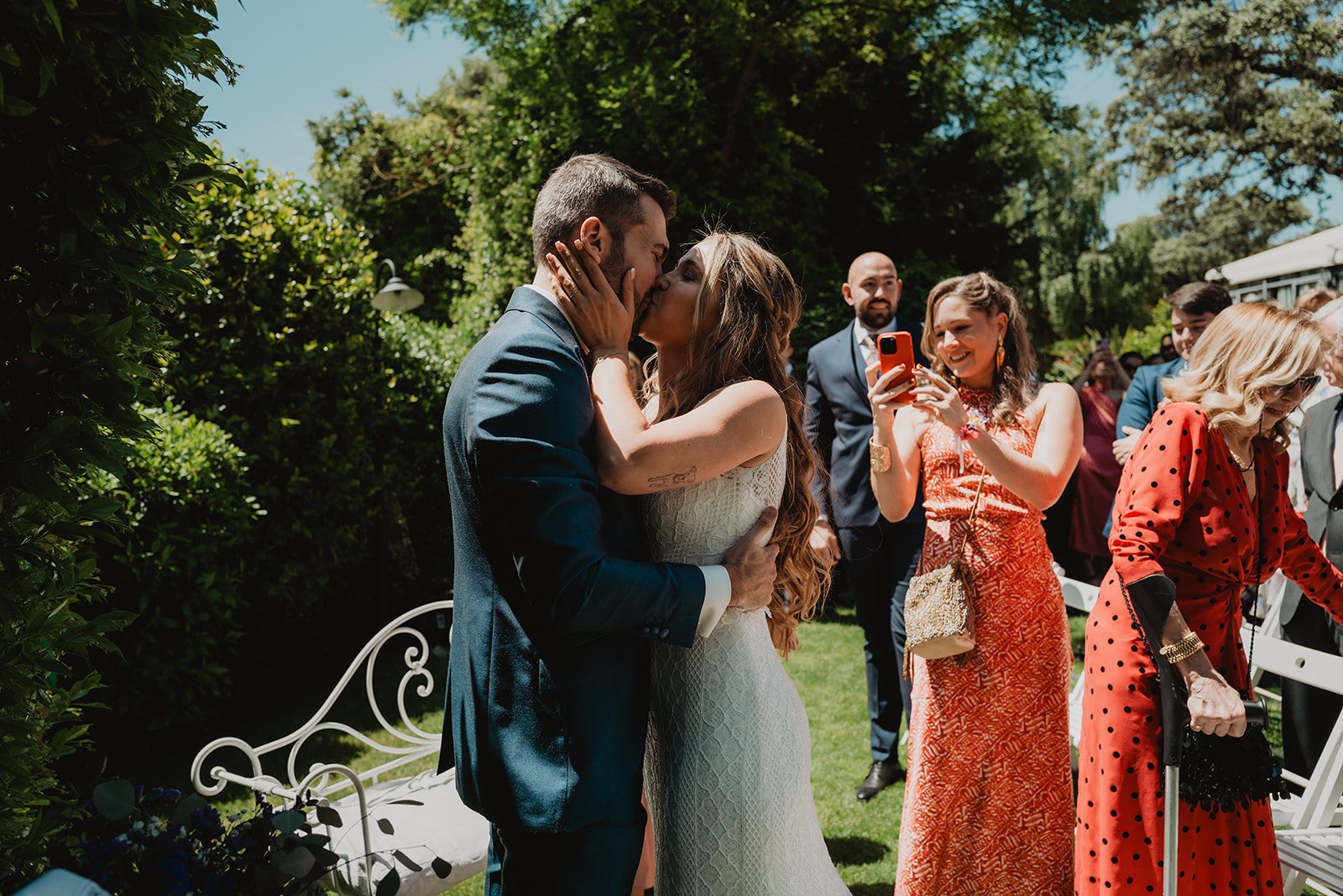 Una pareja besándose en una ceremonia al aire libre con invitados alrededor, algunos tomando fotos. La mujer lleva un vestido blanco y el hombre un traje azul. Los invitados visten ropa elegante.