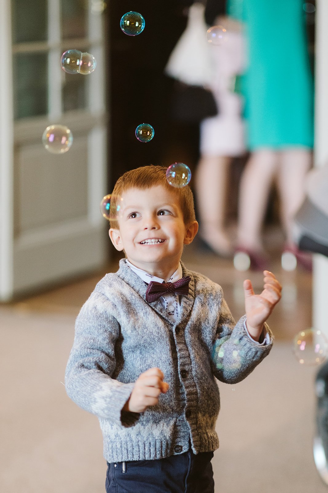Niño sonriente jugando con burbujas, con un suéter gris y pajarita, en un interior.