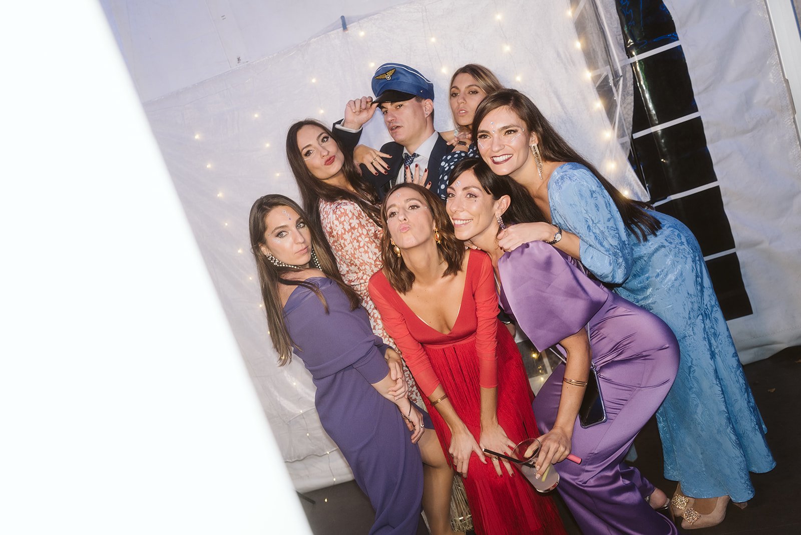 Grupo de personas posando para una foto, algunas haciendo gestos divertidos; las mujeres están vestidas de manera elegante y el hombre lleva un sombrero azul.
