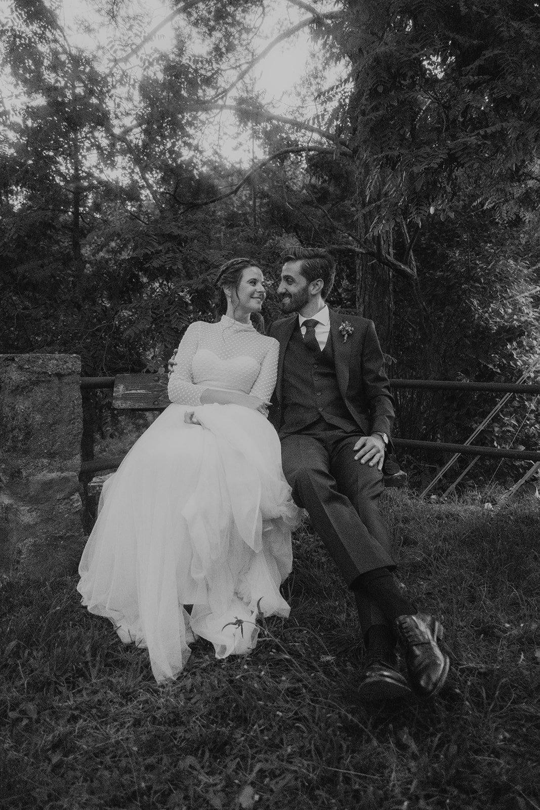 Pareja de recién casados sentada en un banco en un entorno natural, mirándose con sonrisas.