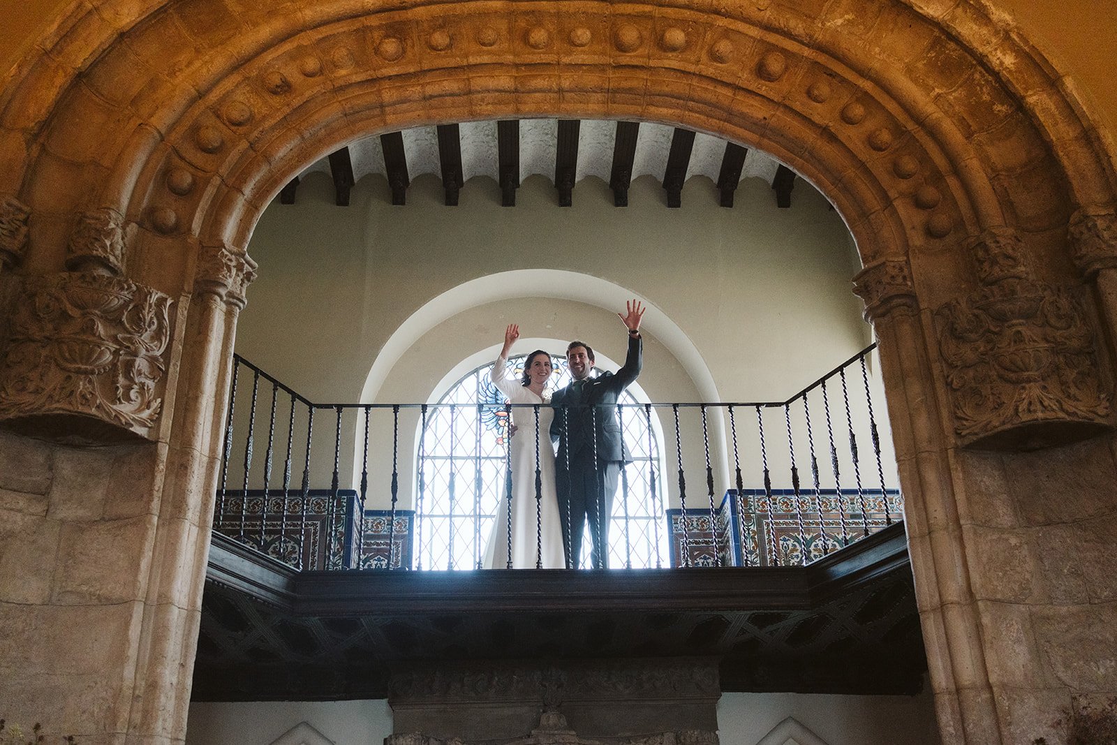 Pareja en un balcón saludando, rodeada de arquitectura histórica con arcos y decoraciones.