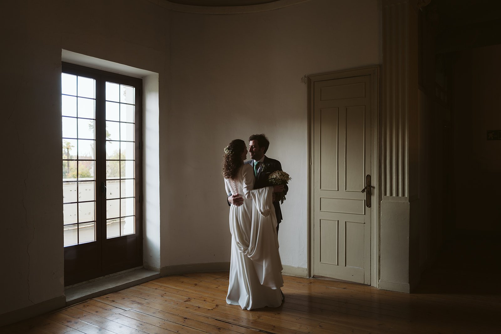 Pareja de novios abrazados en una habitación con luz tenue, ella con vestido de novia y él con traje oscuro, cerca de una puerta de vidrio.
