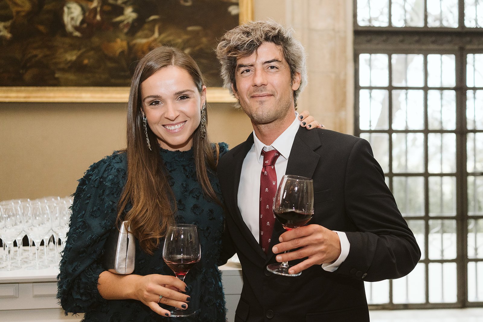 Dos personas en una fiesta, ambos sostienen copas de vino tinto y sonríen, con fondo de ventanas grandes y una pintura.