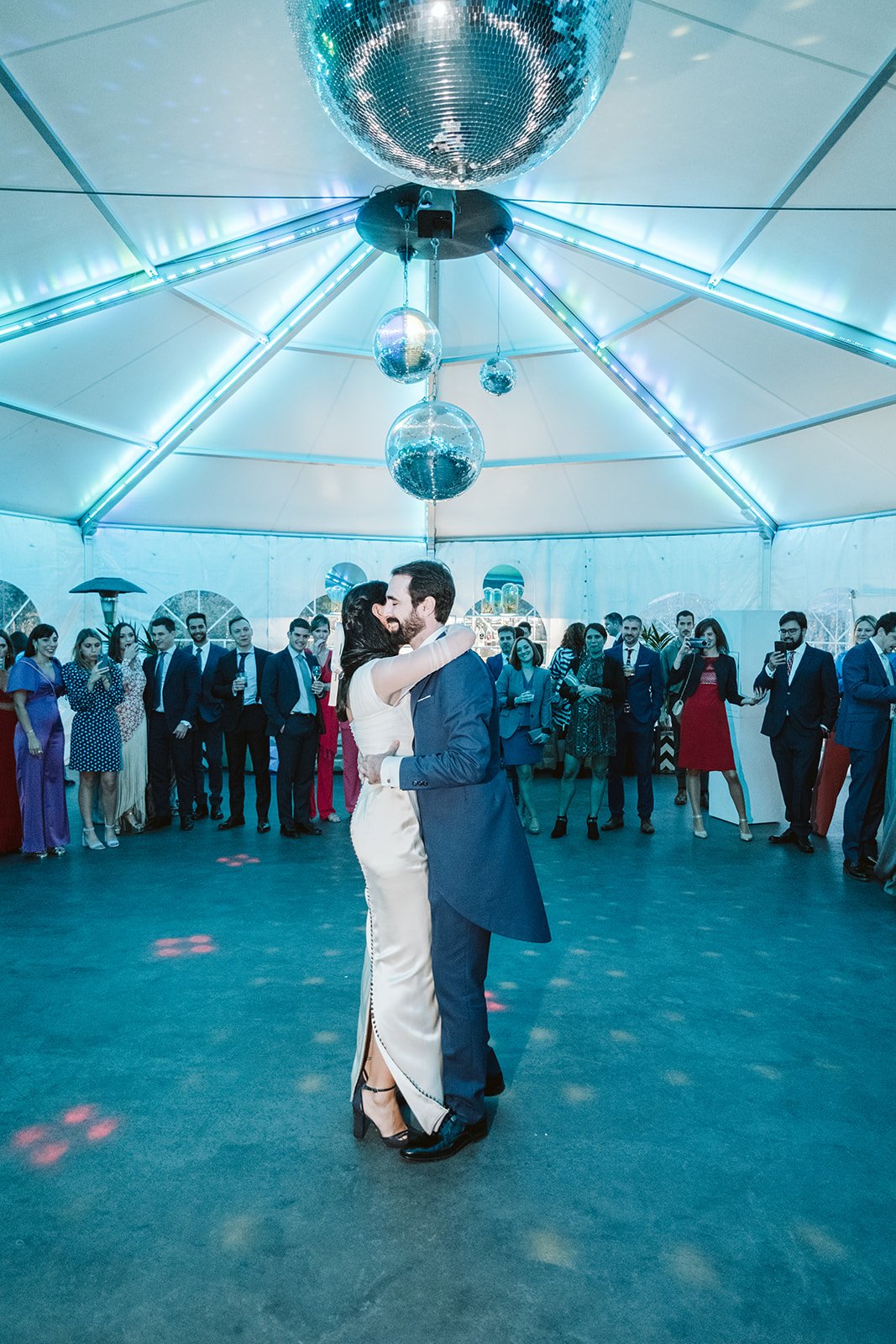 Pareja bailando en una fiesta bajo luces de discoteca, rodeada de invitados vestidos formalmente en una carpa iluminada.