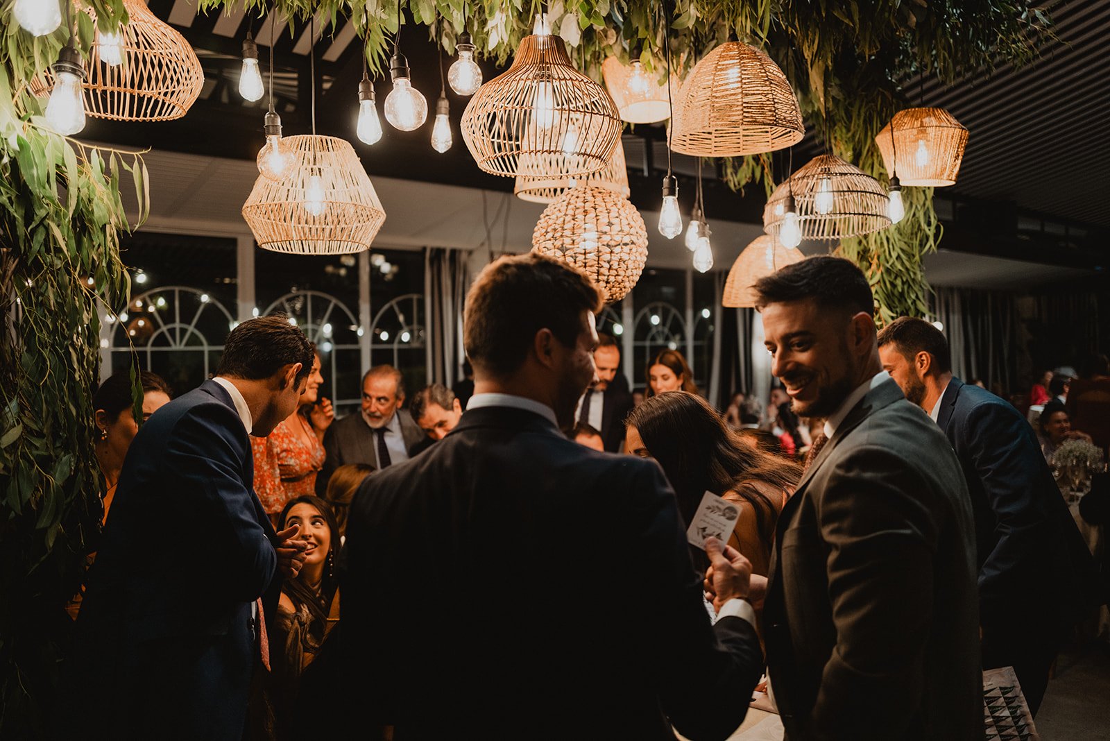 Grupo de personas en un evento social, conversando bajo lámparas colgantes y rodeados de decoración con hojas verdes.