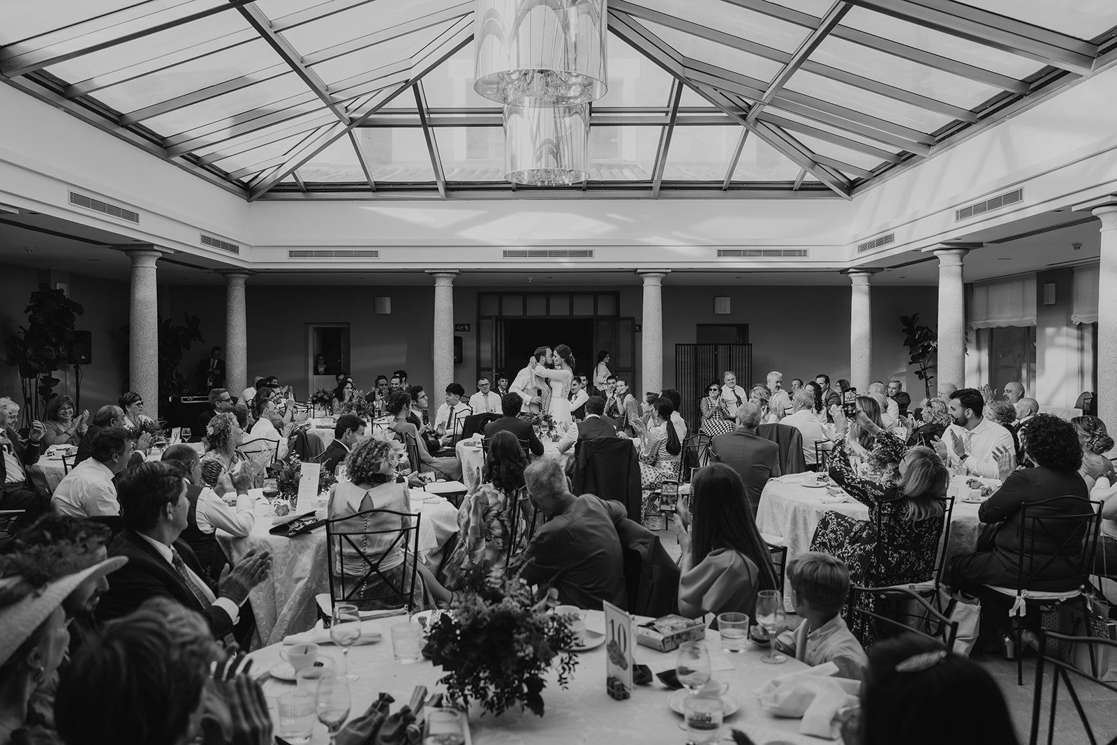 Celebración en un salón con invitados sentados en mesas redondas, bajo un techo de vidrio, presenciando lo que parece ser una boda o evento formal.