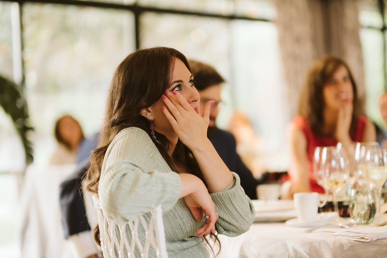 Mujer sentada en una mesa, apoyando su mano en la cara, con expresión pensativa. Al fondo, varias personas en ambiente de evento social.