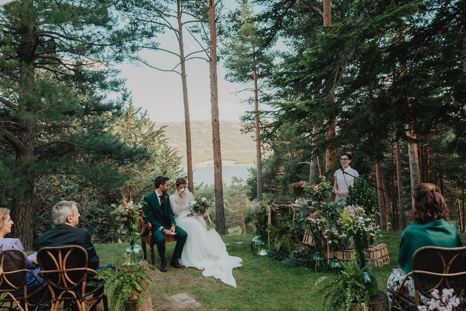 Boda al aire libre en un bosque, con una pareja de novios sentados, rodeados de invitados y decoraciones florales. Un oficiante está de pie hablando.