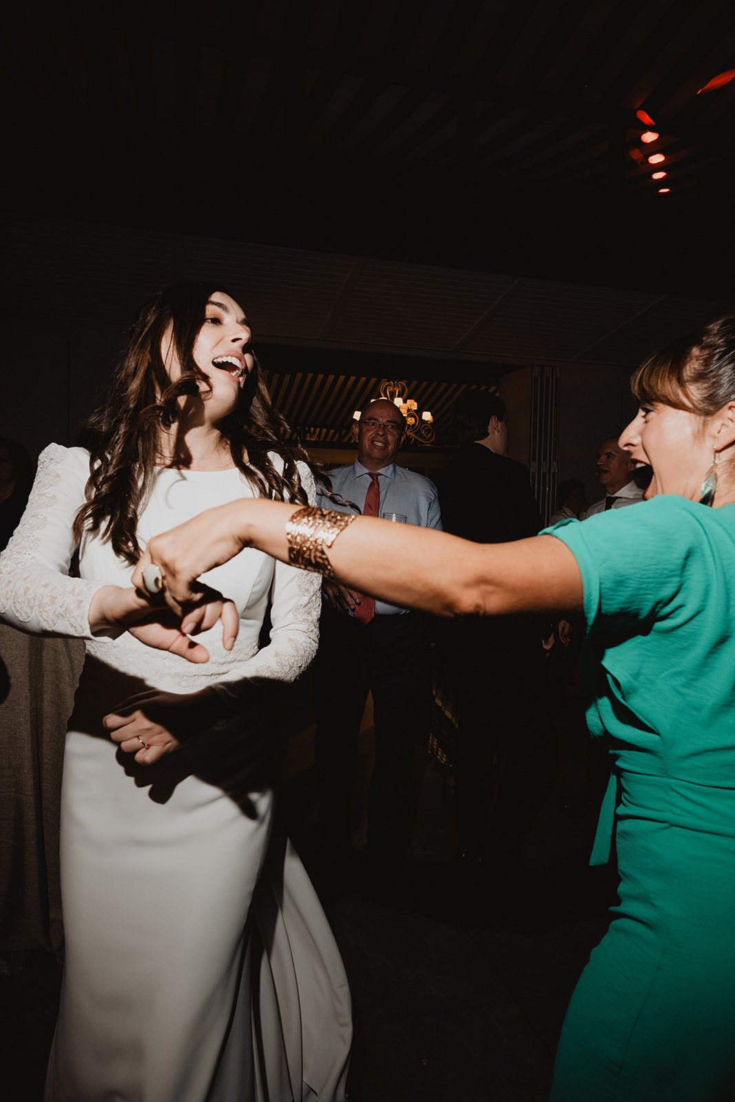Dos mujeres bailando en una fiesta, una vestida de blanco y otra de verde, con personas observando en el fondo.
