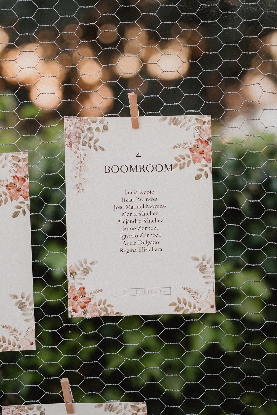 Panel decorativo colgado en una malla, con una lista de nombres para un evento, adornado con motivos florales.