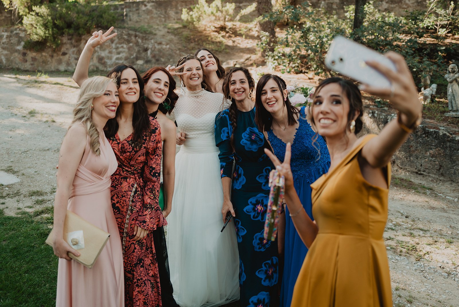 Un grupo de mujeres sonríe mientras se toman una selfie en una boda, con vestidos coloridos y elegantes, rodeadas de un entorno natural.