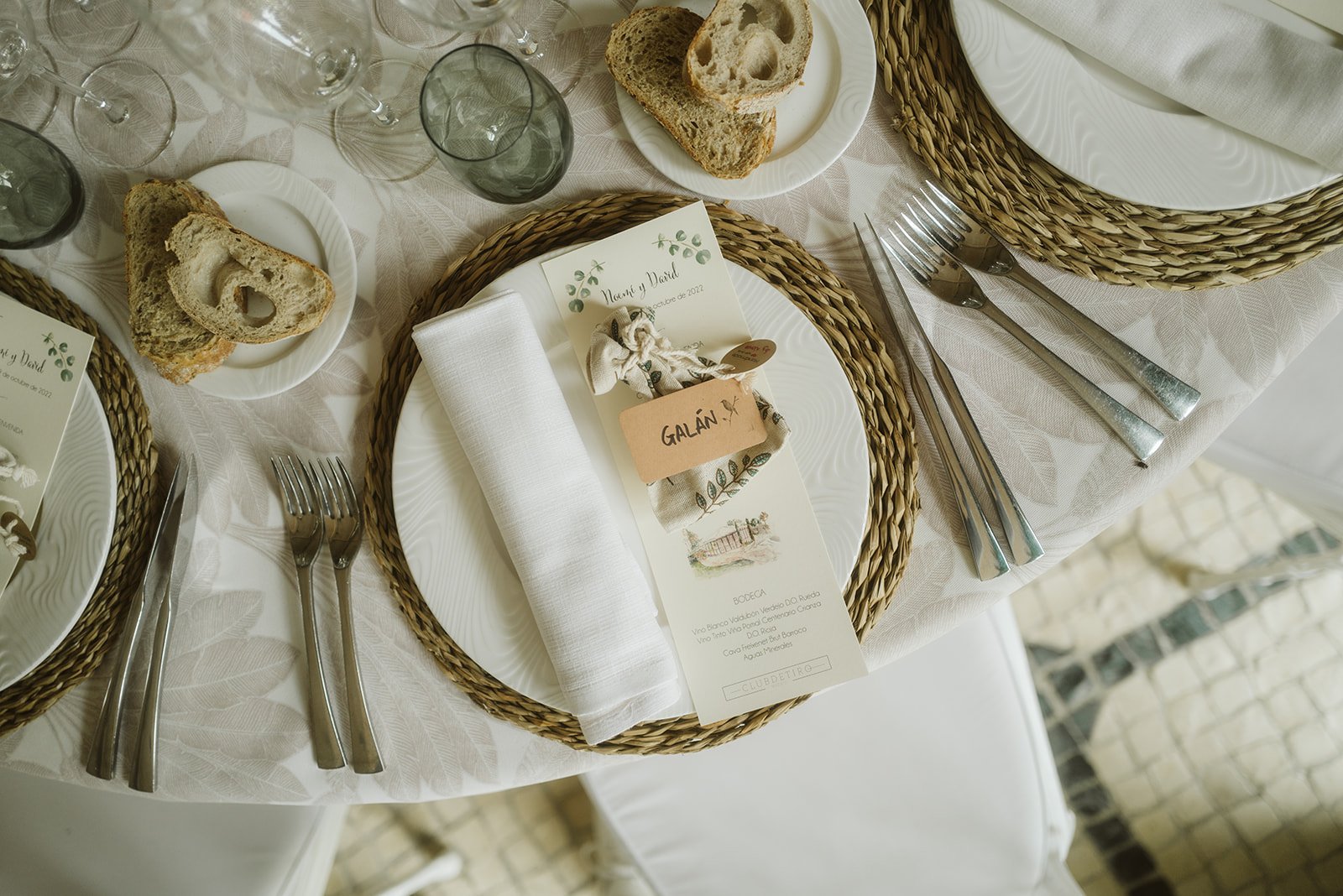 Mesa elegantemente puesta con platos de cerámica blanca, cubiertos de acero inoxidable, servilletas blancas, vasos de cristal y rebanadas de pan. Hay tarjetas de menú decorativas y una tarjeta con el nombre "Galán".