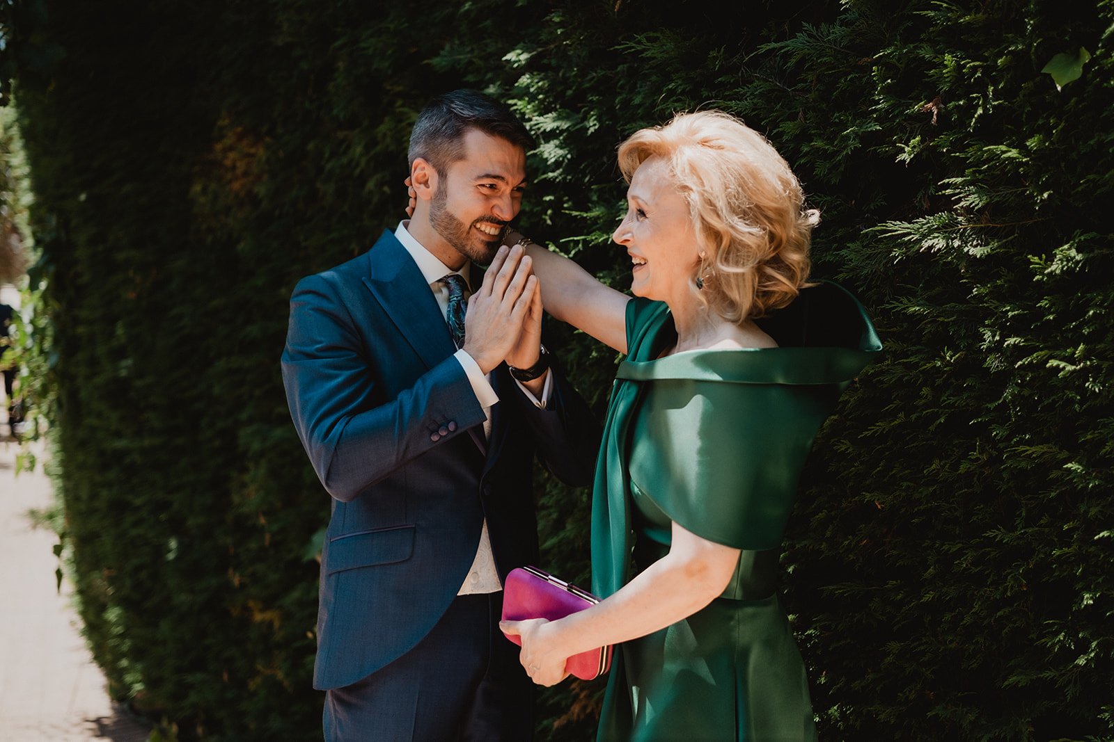 Hombre y mujer elegante sonriendo frente a un seto, el hombre lleva un traje formal y la mujer un vestido verde, sosteniendo un bolso morado.