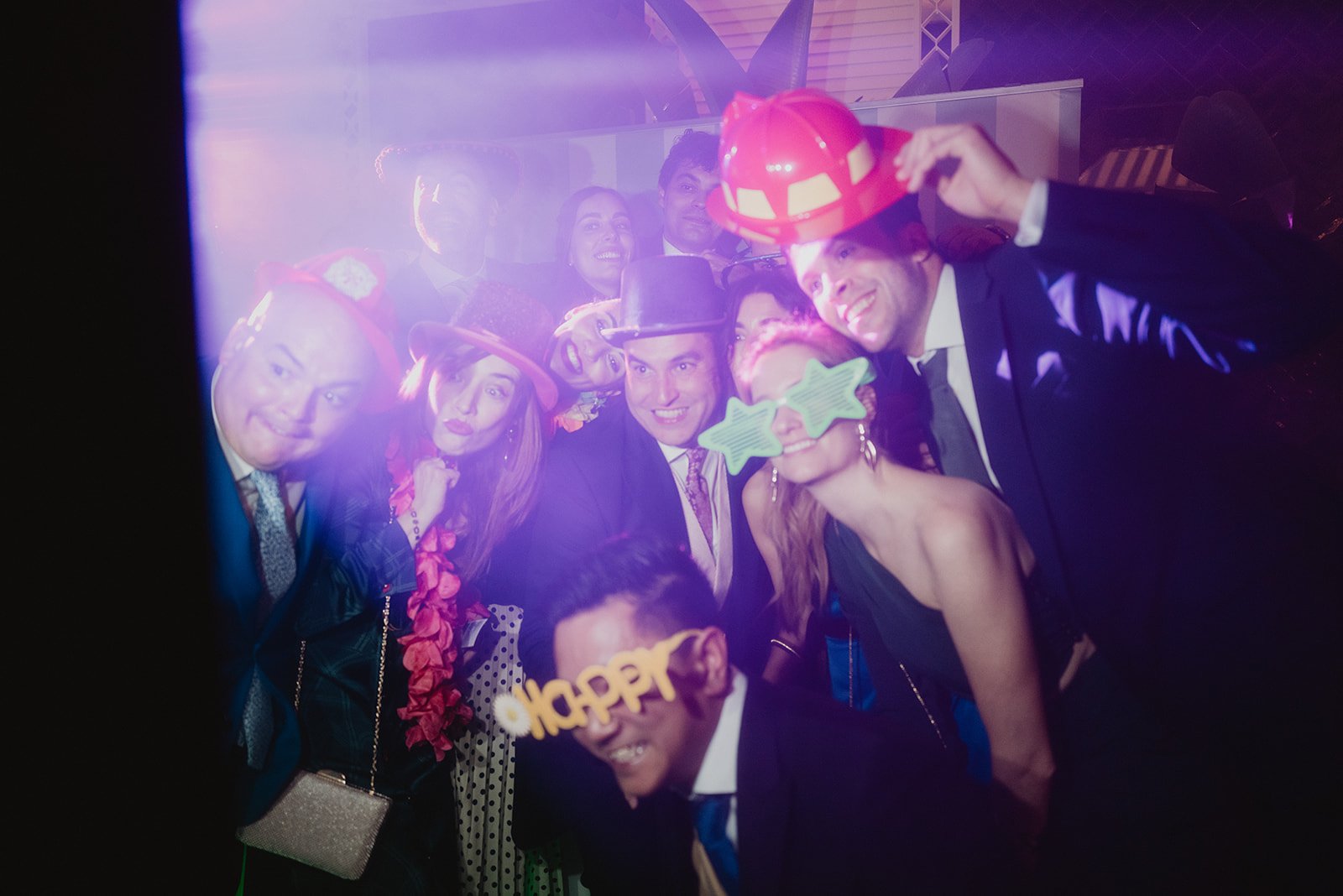 Grupo de personas en una fiesta, vistiendo trajes formales y accesorios divertidos como sombreros y gafas de fiesta. Hay iluminación de colores y un ambiente alegre.