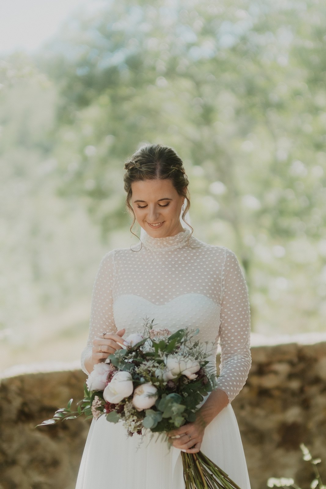 Novia con vestido blanco de encaje sosteniendo un ramo de flores, fondo natural borroso.