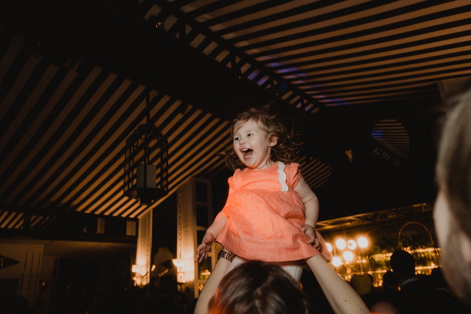 Una niña pequeña con vestido naranja siendo levantada en el aire, sonriendo en un ambiente interior con techo a rayas y luces cálidas.