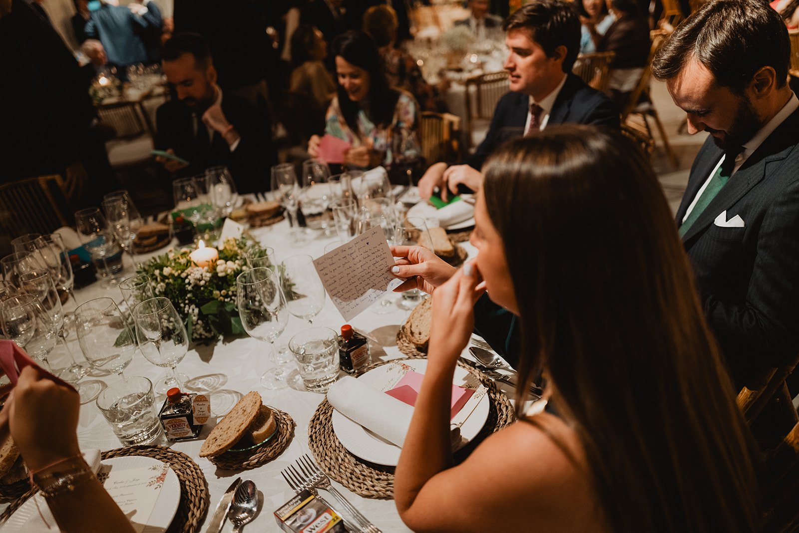 Personas sentadas en una mesa elegante durante un evento, con copas de vino, velas y platos servidos con pan. Una persona sostiene una tarjeta en la mano.