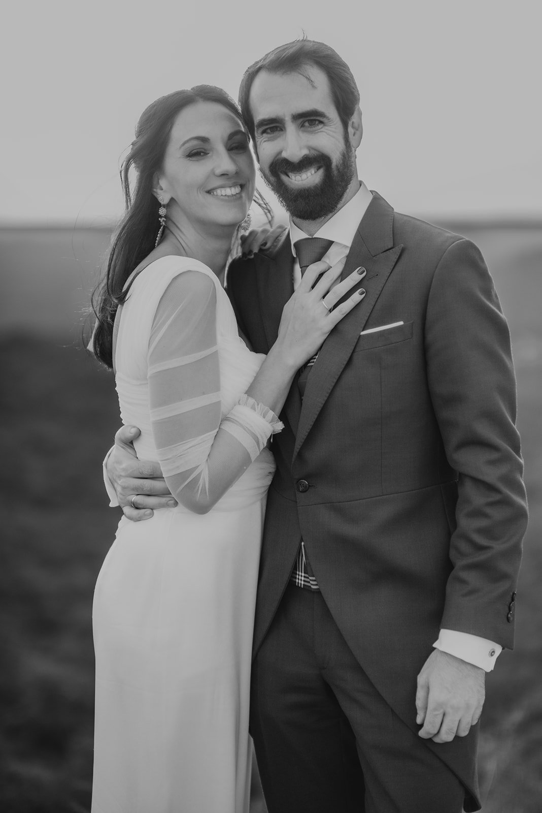 Pareja sonriente vestida de novios, ella con vestido de boda y él con traje formal, abrazados y felices al aire libre.