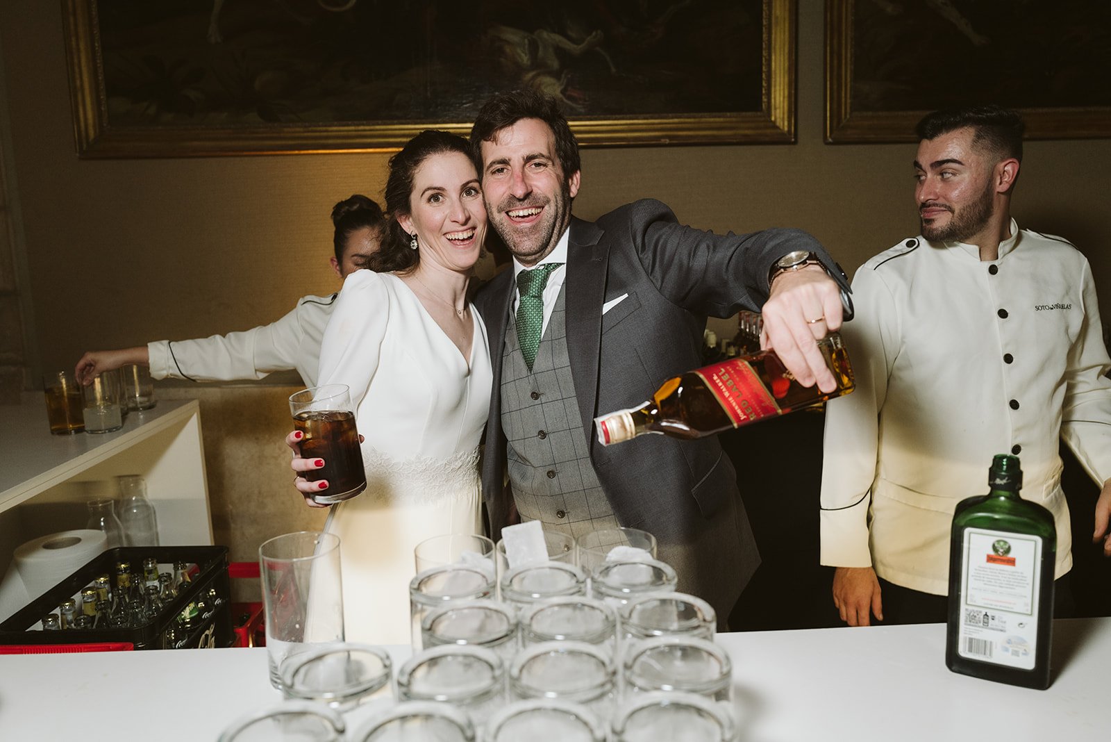 Una pareja sonriente en una celebración, con la mujer vistiendo un vestido blanco y el hombre un traje elegante. Él está sirviendo whisky Johnnie Walker mientras un bartender observa. Hay varios vasos y una botella de Jägermeister en la barra.