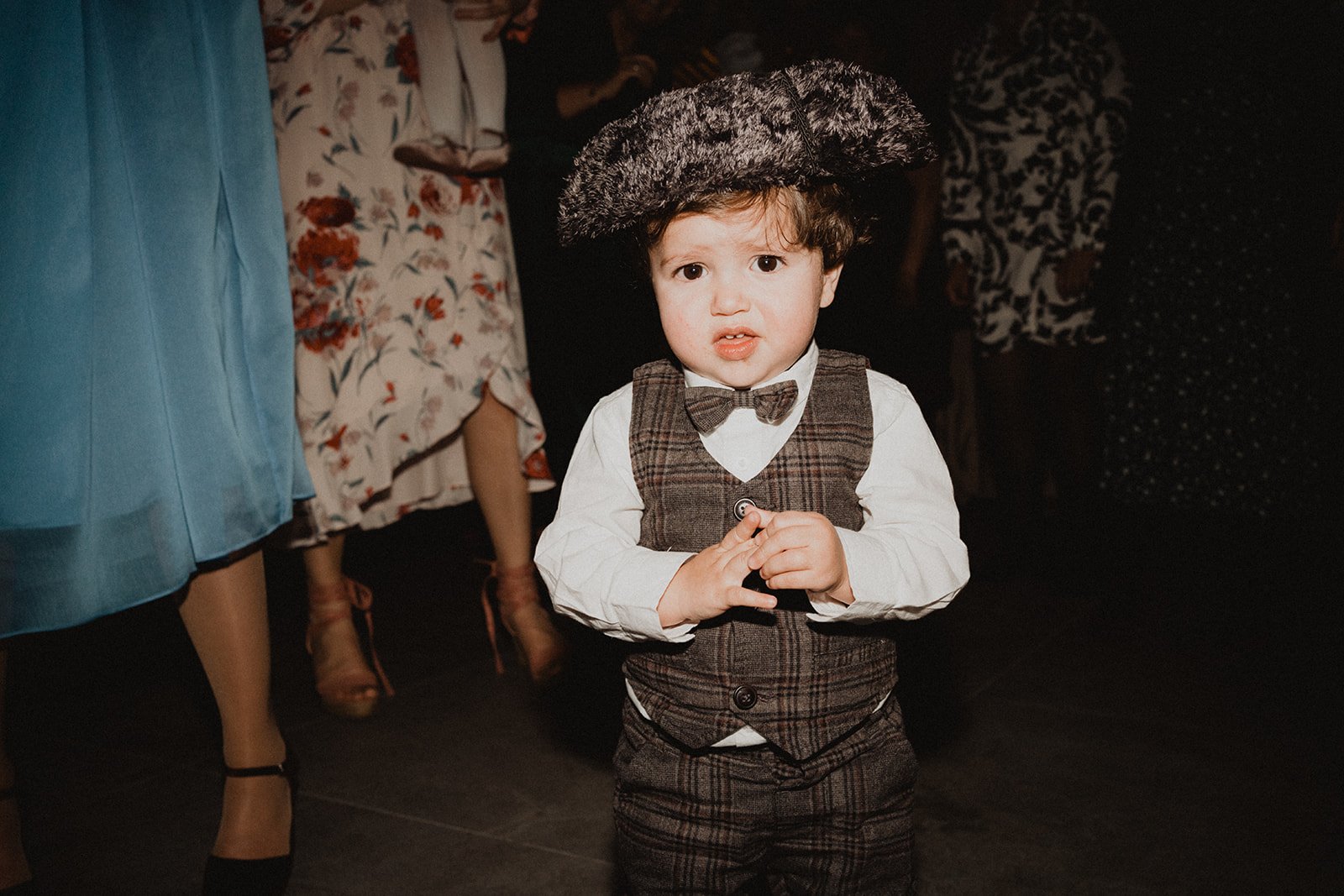 Niño pequeño con traje elegante y boina, rodeado de personas vestidas formalmente en un evento.