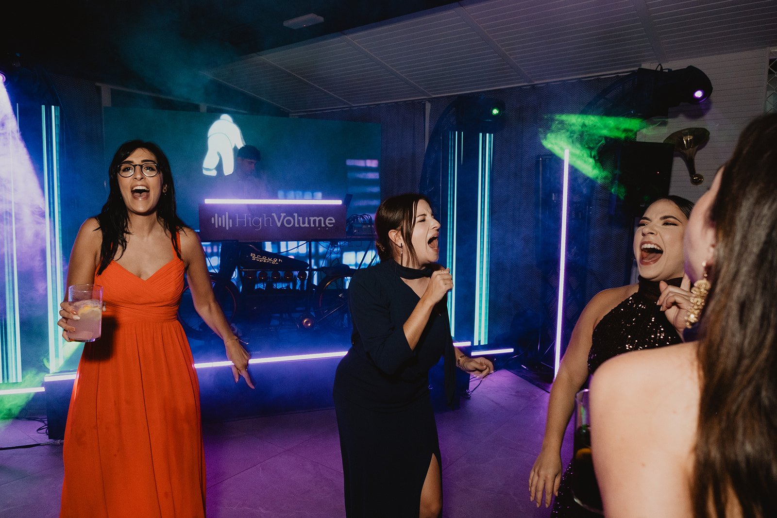 Personas divirtiéndose y bailando en una fiesta con luces coloridas y un DJ al fondo.