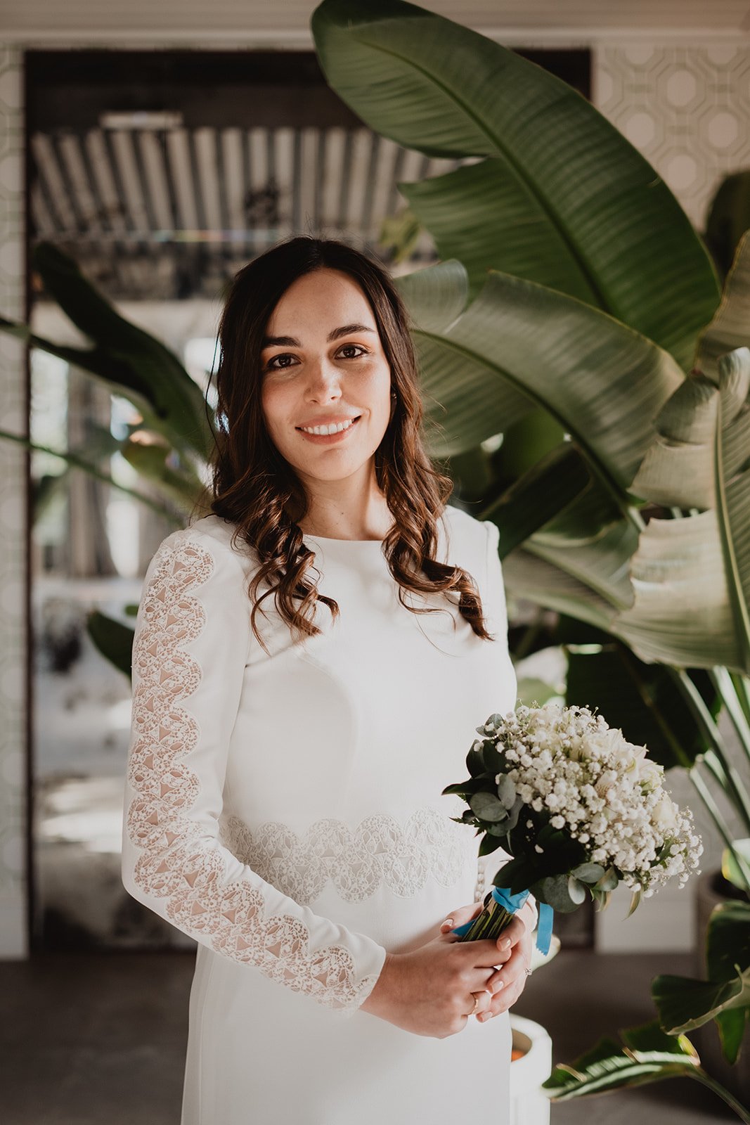 Mujer con vestido blanco sosteniendo ramo de flores, rodeada de plantas verdes.