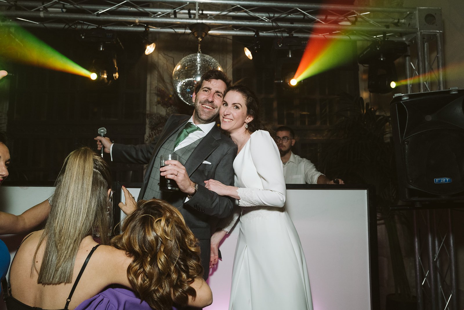 Pareja vestida formalmente sonriendo en una fiesta con luces de discoteca y una bola de espejos en el fondo.