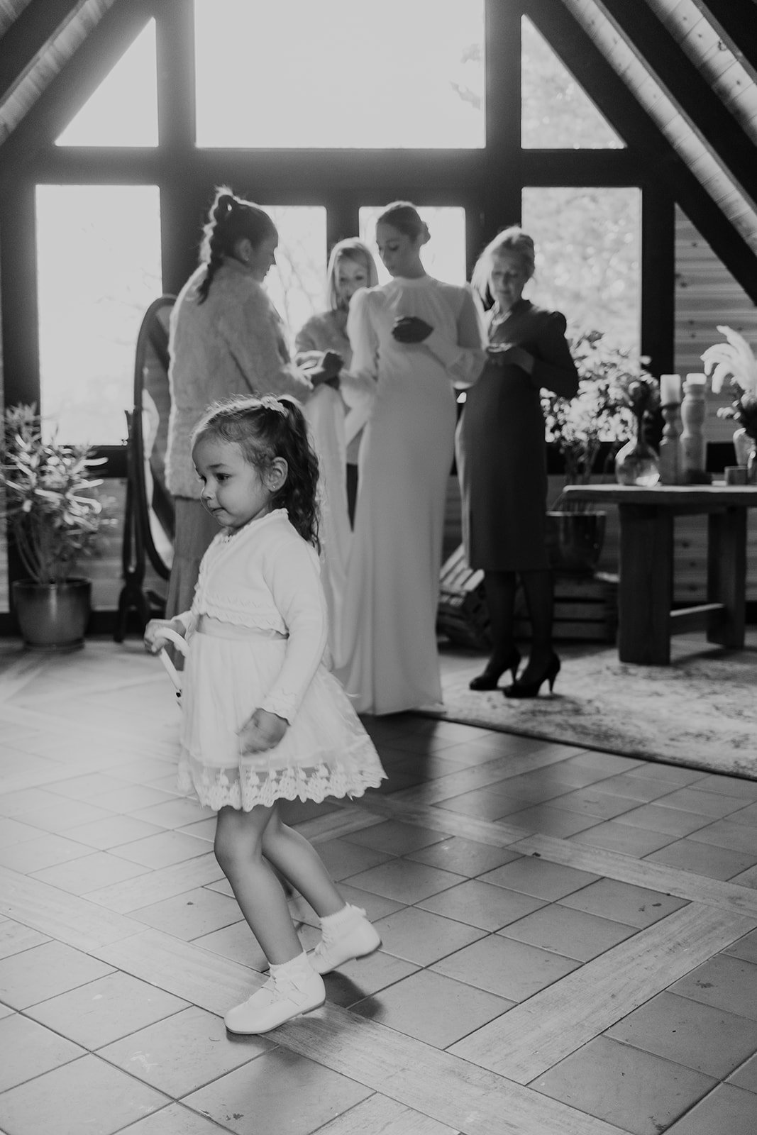 Una niña pequeña con vestido blanco camina en un interior, mientras al fondo cuatro mujeres están reunidas, dos de ellas con vestidos largos posiblemente de boda. La escena es en blanco y negro.