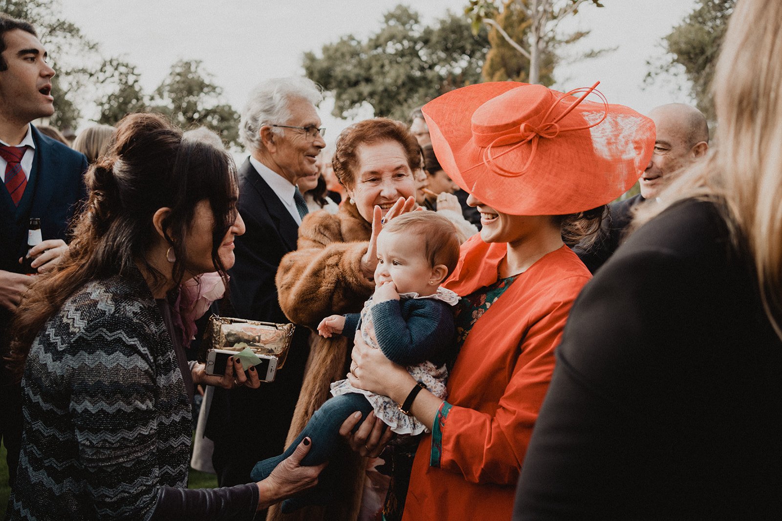 Grupo de personas en un evento, alguna sostiene a un bebé, otra lleva un sombrero rojo elegante, y están conversando y sonriendo.