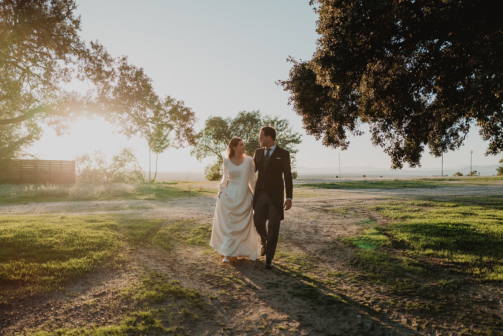Una pareja vestida de novios caminando juntos en un entorno natural con árboles y luz de atardecer.