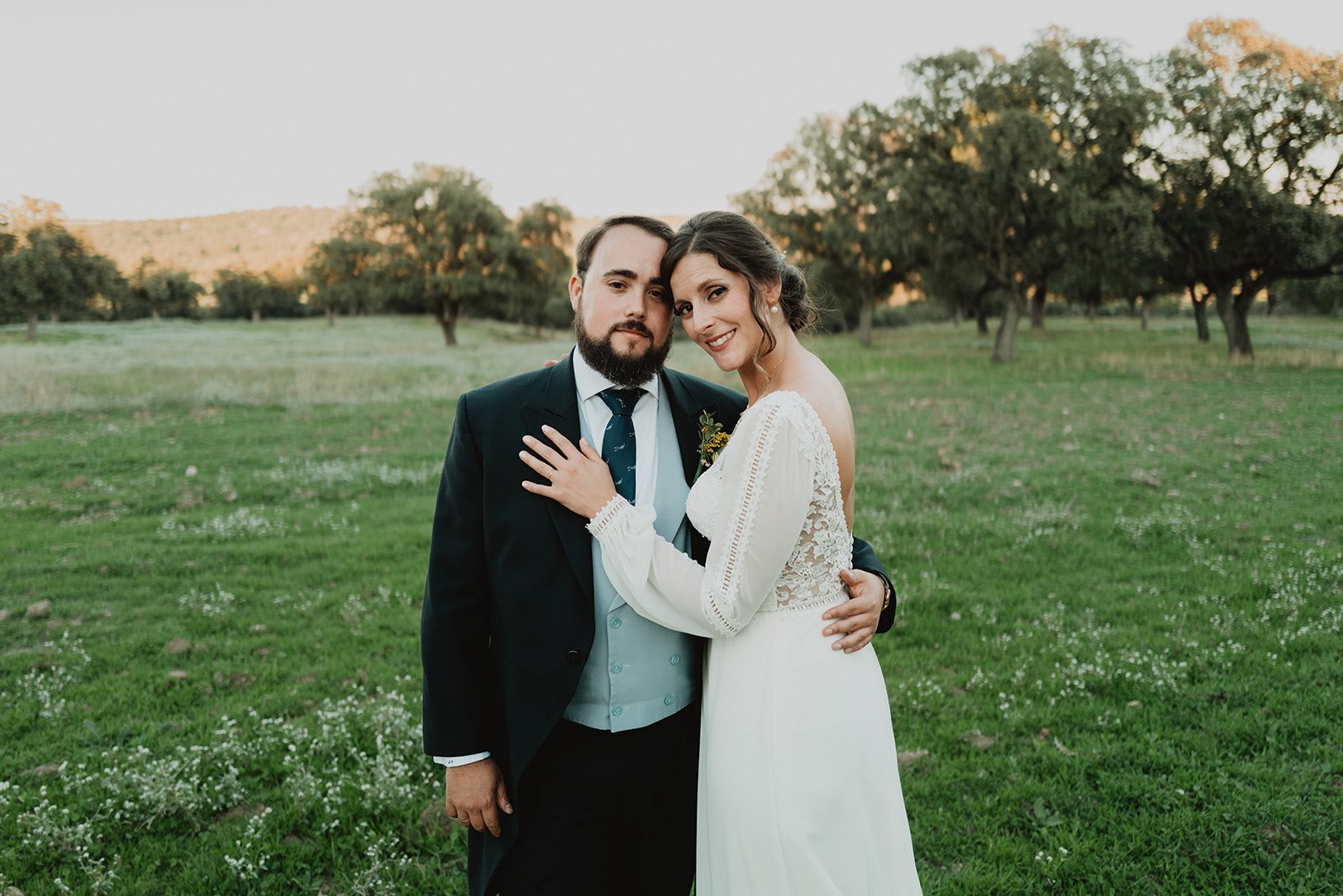 Pareja de recién casados posando en un campo verde con árboles al fondo.