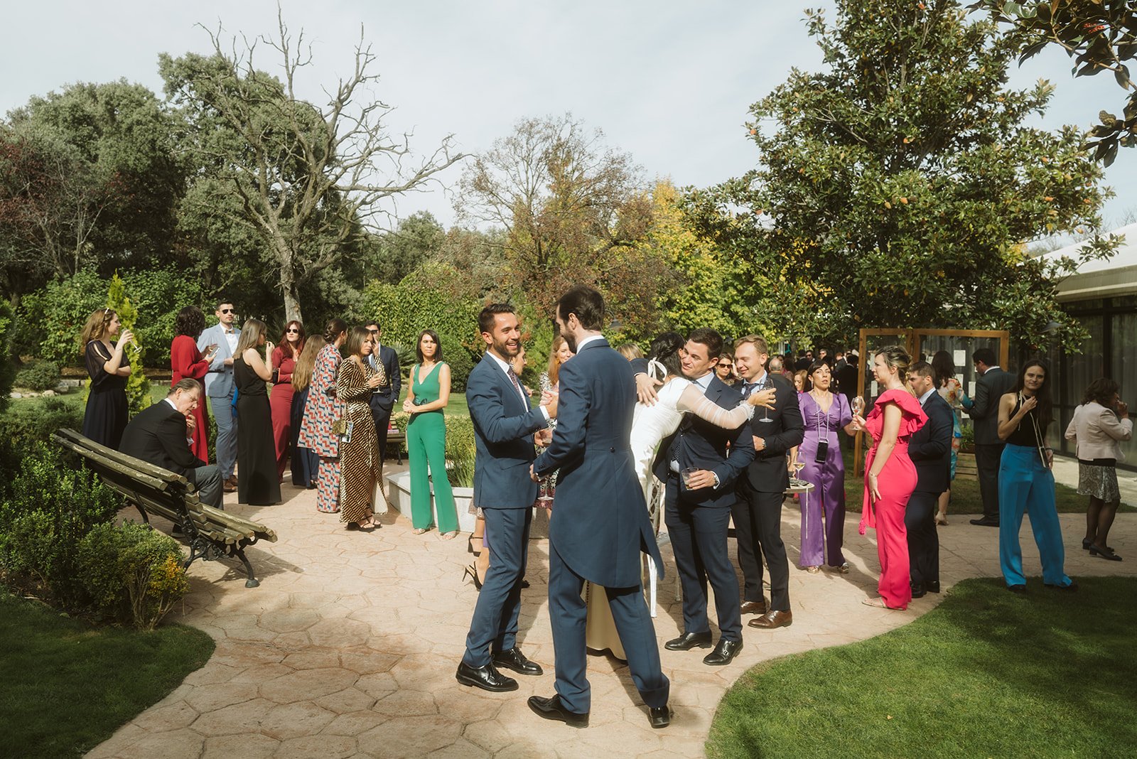 Grupo de personas vestidas de gala reunidas en un evento al aire libre, abrazándose y conversando en un jardín.