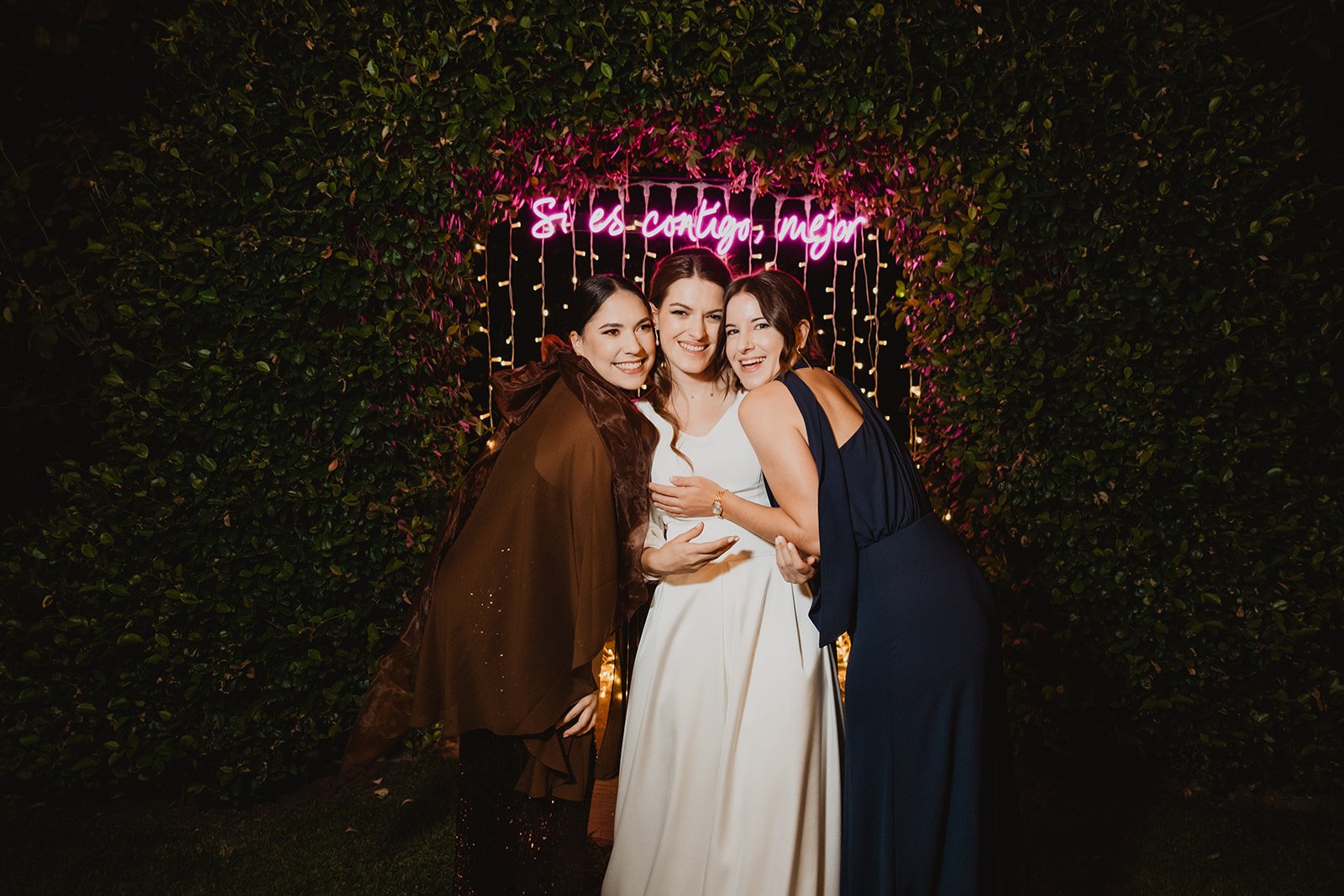 Tres mujeres sonrientes posan frente a un letrero de neón rosa que dice ‘Si es contigo, mejor’, rodeadas de luces y vegetación.