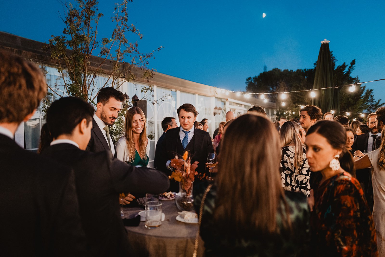 Personas en un evento social al aire libre por la noche, vestidas formalmente, conversando alrededor de una mesa con bebidas y decoraciones, bajo luces colgantes.