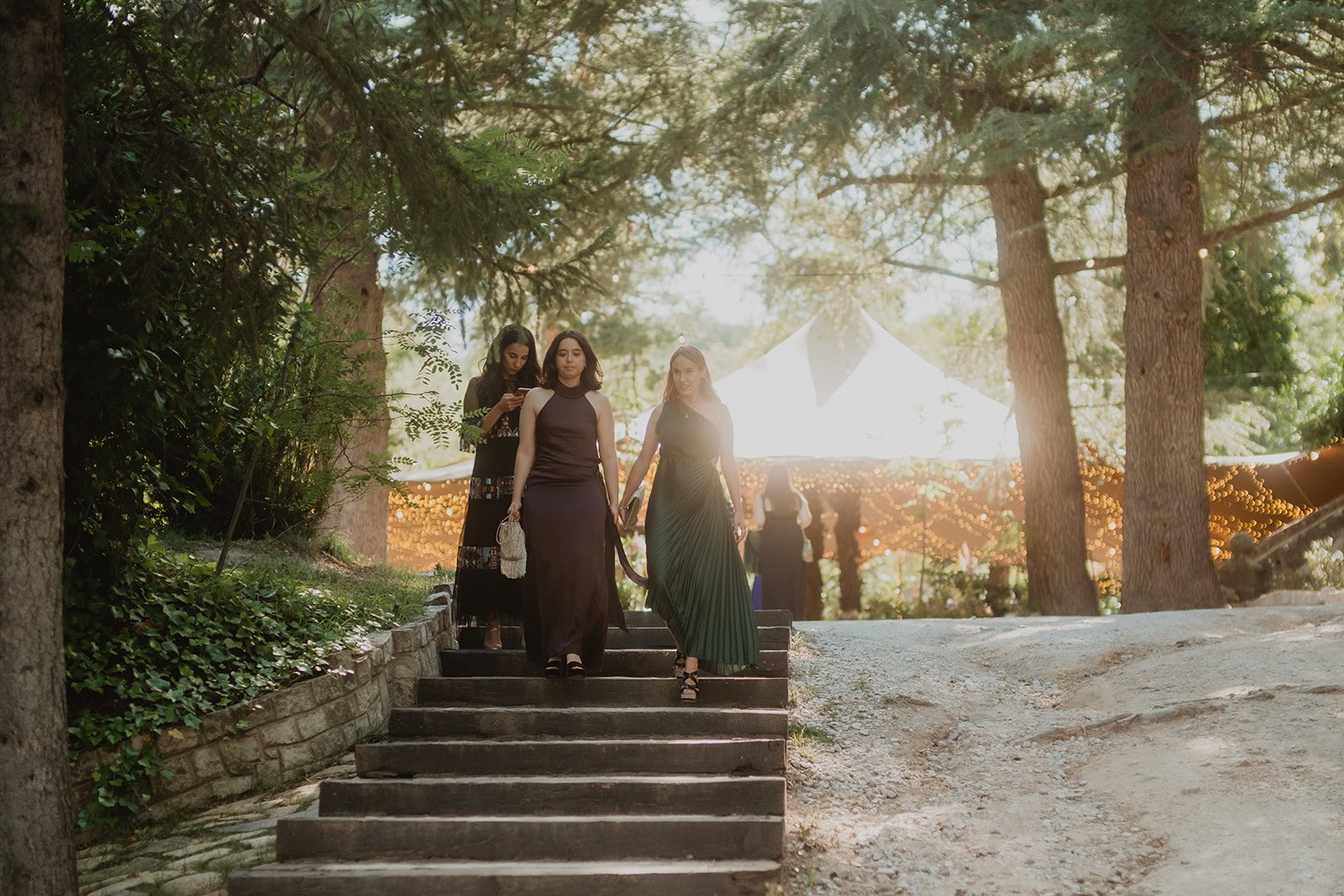 Mujeres bajando escaleras en un entorno al aire libre con árboles y luces decorativas, llevando vestidos formales.