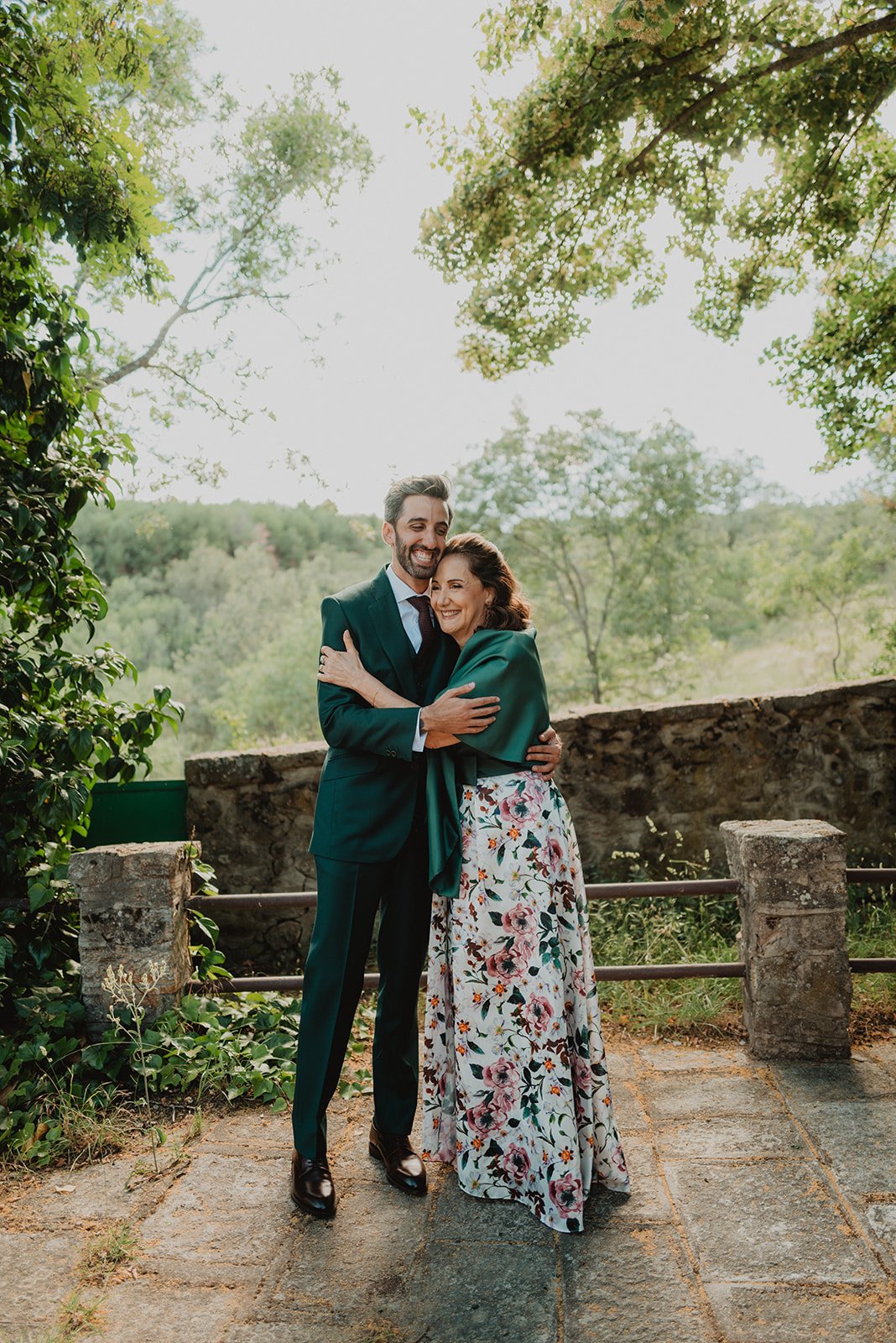 Pareja abrazándose al aire libre, rodeada de árboles y naturaleza. Él viste traje verde y ella un vestido floral con chal verde.