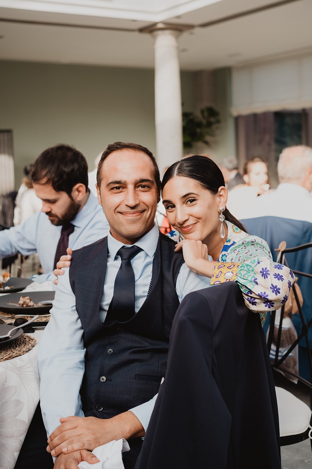 Pareja sonriendo en una mesa durante un evento, rodeados de otras personas.